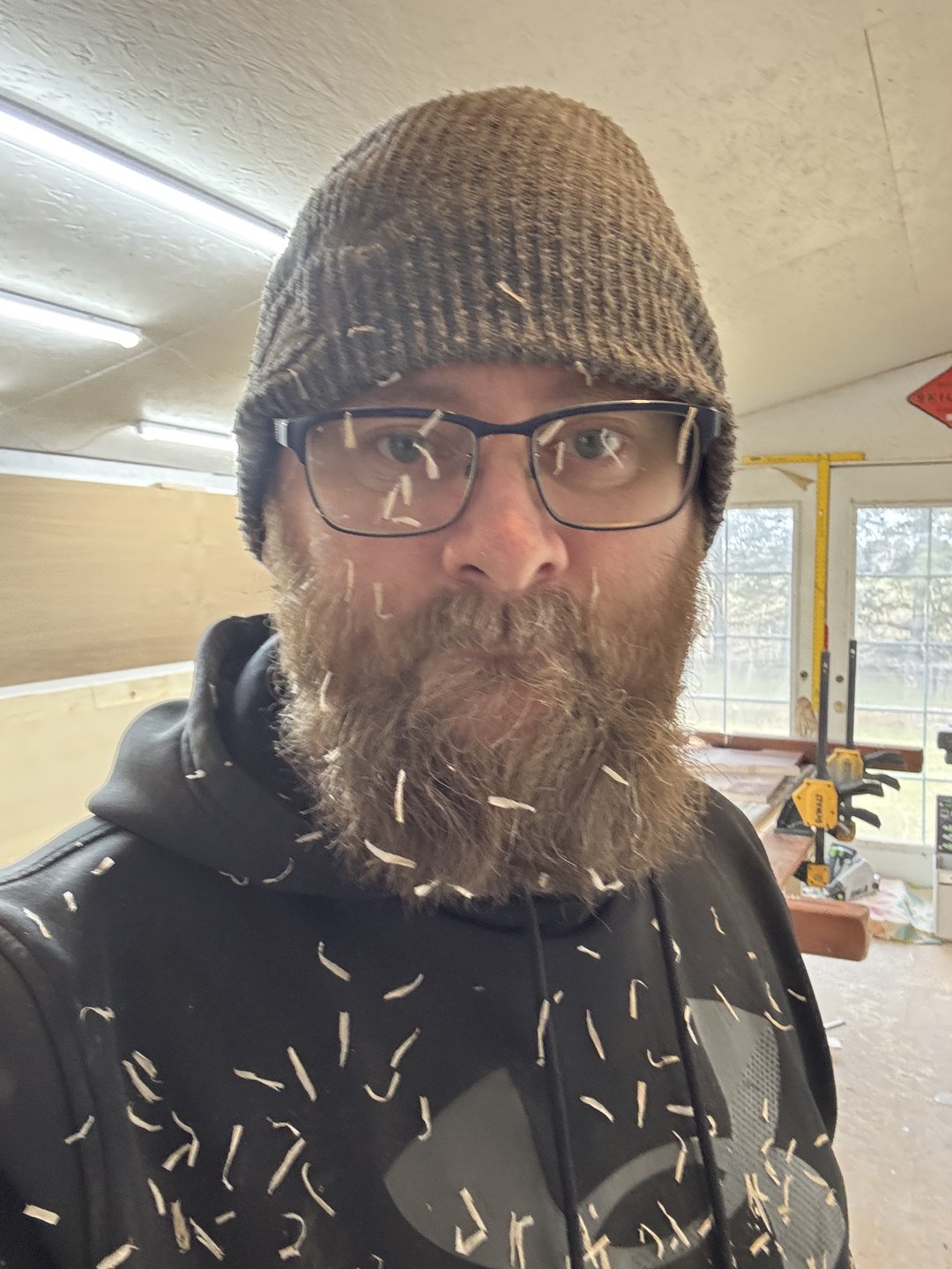 Ben Rawson in his woodworking shop, wearing a knitted hat and glasses, with wood shavings on his hoodie
